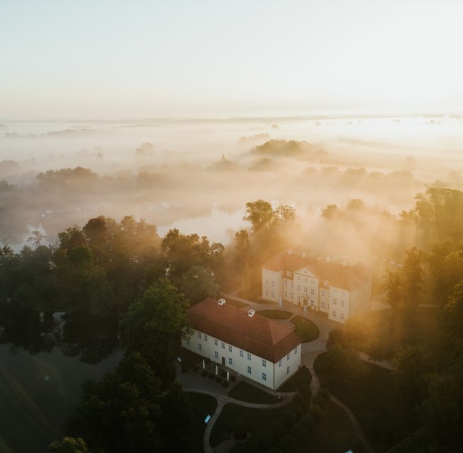 Goldenes Licht taucht Schloss Mirow in eine mystische Atmosph&auml;re. Nebelschwaden ziehen &uuml;ber die Seenlandschaft der Mecklenburgischen Seenplatte und lassen das historische Ensemble aus dem 18. Jahrhundert wie aus der Zeit gefallen wirken. Ein Moment purer Stille zwischen Wasser und Wald., &copy; 1000seen.de