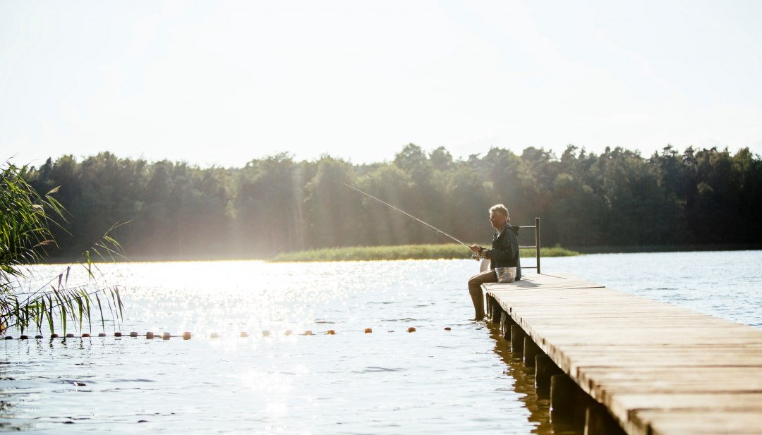 Ein Mann sitzt auf einem Holzsteg an einem ruhigen See und angelt, während die Sonne das Wasser glitzern lässt.