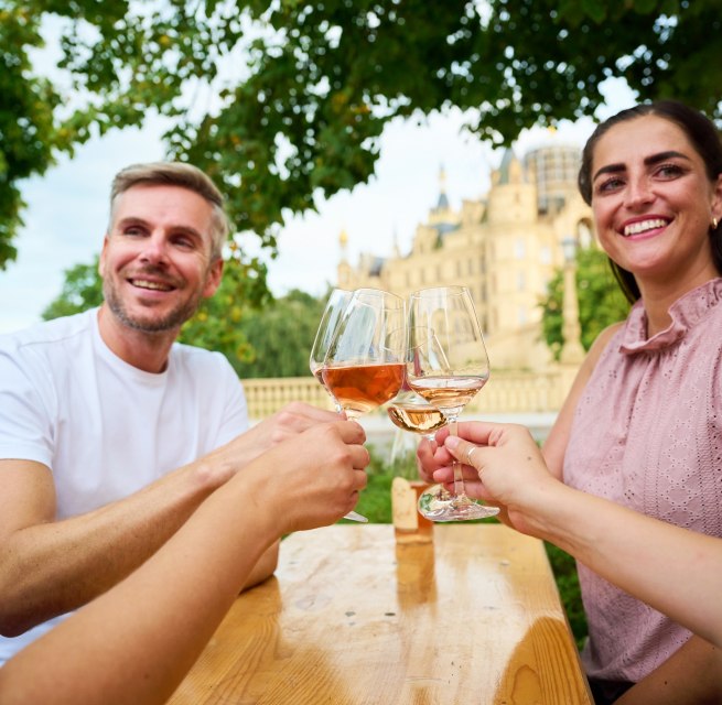 Gasten proosten met een glas wijn tijdens het wijnfeest in Schwerin op de Alte Garten. Op de achtergrond doemt het Slot Schwerin op en vormt zo het bijzondere decor voor het wijnfeest. // &copy; LHSN/Celien Graubaum