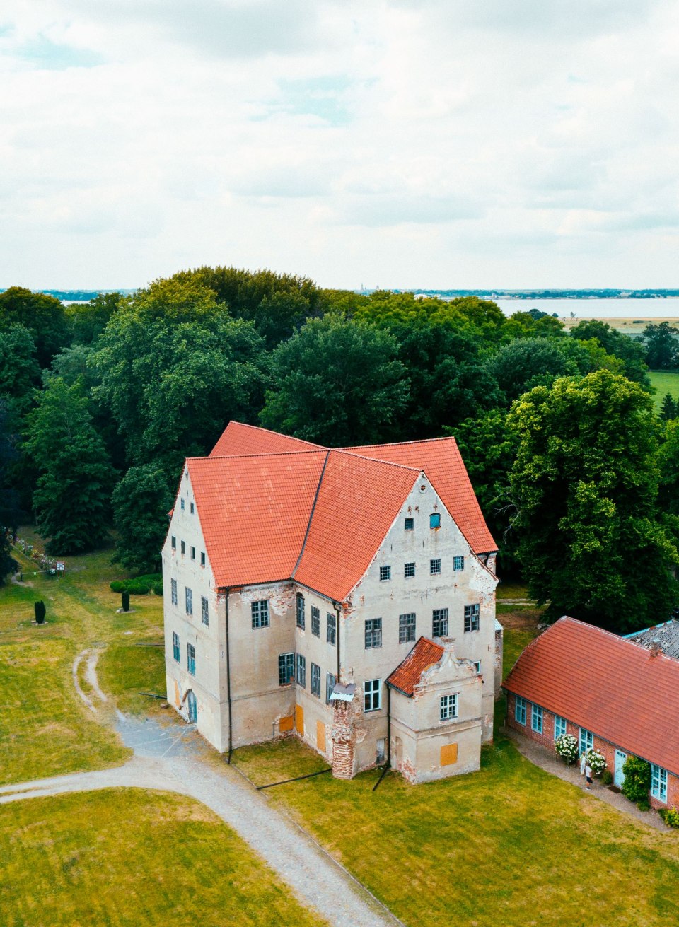 Luftaufnahme eines hellen Schlosses mit roten Dächern zwischen Bäumen, dahinter glänzt das Wasser am Horizont. // Aus der Luft zeigt sich das Schloss Ludwigsburg zwischen alten Bäumen. Dahinter öffnet sich der Blick auf das Wasser am Horizont, und weite Wiesen laden zum Verweilen ein. // © MV-T/Friedrich Luftaufnahme eines hellen Schlosses mit roten Dächern zwischen Bäumen, dahinter glänzt das Wasser am Horizont.