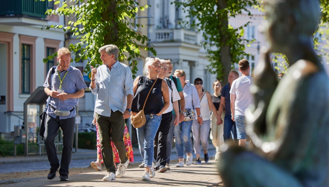Eine Gruppe Menschen ist mit einem Stadtführer auf einem gepflasterten Weg unterwegs. Rechts, unscharf im Bild ist eine Statue am Pfaffenteich zu sehen, im Hintergrund erscheinen die Villen. Blickpunkt sind der Stadtführer sowie ein Paar, dass sich angeregt unterhält., © Oliver Borchert Eine Gruppe Menschen ist mit einem Stadtführer auf einem gepflasterten Weg unterwegs. Rechts, unscharf im Bild ist eine Statue am Pfaffenteich zu sehen, im Hintergrund erscheinen die Villen. Blickpunkt sind der Stadtführer sowie ein Paar, dass sich angeregt unterhält., © Oliver Borchert