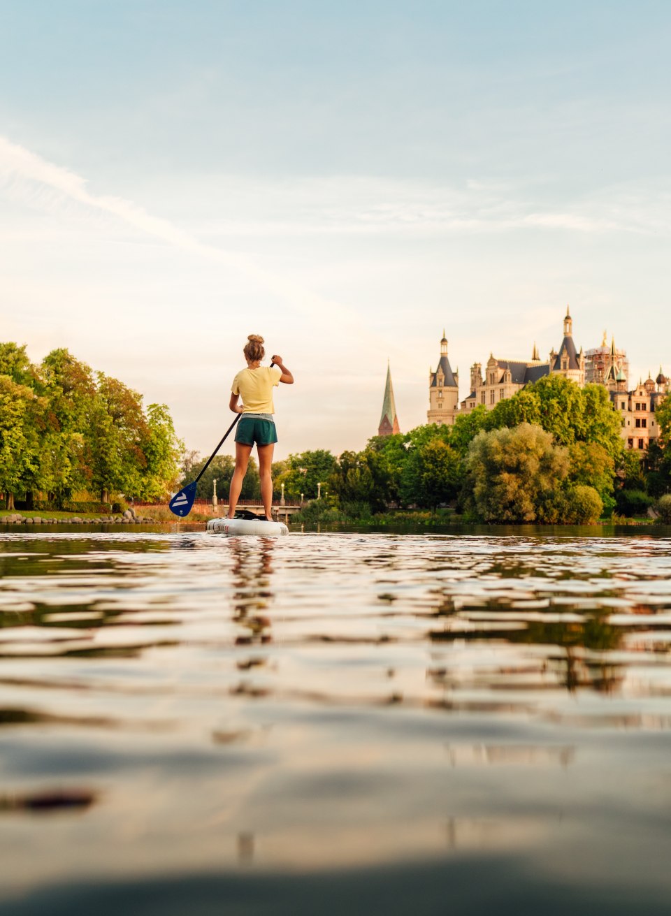 Eine Person steht beim Stand-up-Paddling auf dem Schweriner See und paddelt im warmen Abendlicht auf das Schloss Schwerin zu.
