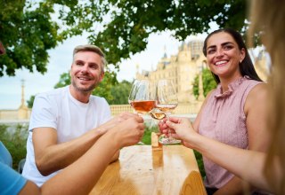 Gasten proosten met een glas wijn tijdens het wijnfeest in Schwerin op de Alte Garten. Op de achtergrond doemt het Slot Schwerin op en vormt zo het bijzondere decor voor het wijnfeest. // © LHSN/Celien Graubaum Gasten proosten met een glas wijn tijdens het wijnfeest in Schwerin op de Alte Garten. Op de achtergrond doemt het Slot Schwerin op en vormt zo het bijzondere decor voor het wijnfeest. // © LHSN/Celien Graubaum