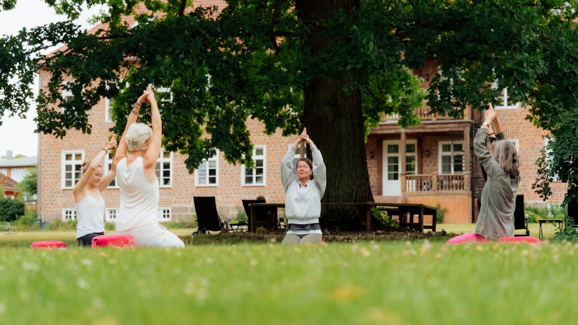 Drei Frauen machen Yoga auf einer Wiese unter einem großen Baum, im Hintergrund steht das Gutshaus Ludorf. // Unter einer alten Eiche fließen ruhige Bewegungen durch den Gutshauspark Ludorf – Yoga auf der Wiese, Backstein im Blick, Kopf wird frei // © MV-T/Petermann Drei Frauen machen Yoga auf einer Wiese unter einem großen Baum, im Hintergrund steht das Gutshaus Ludorf.