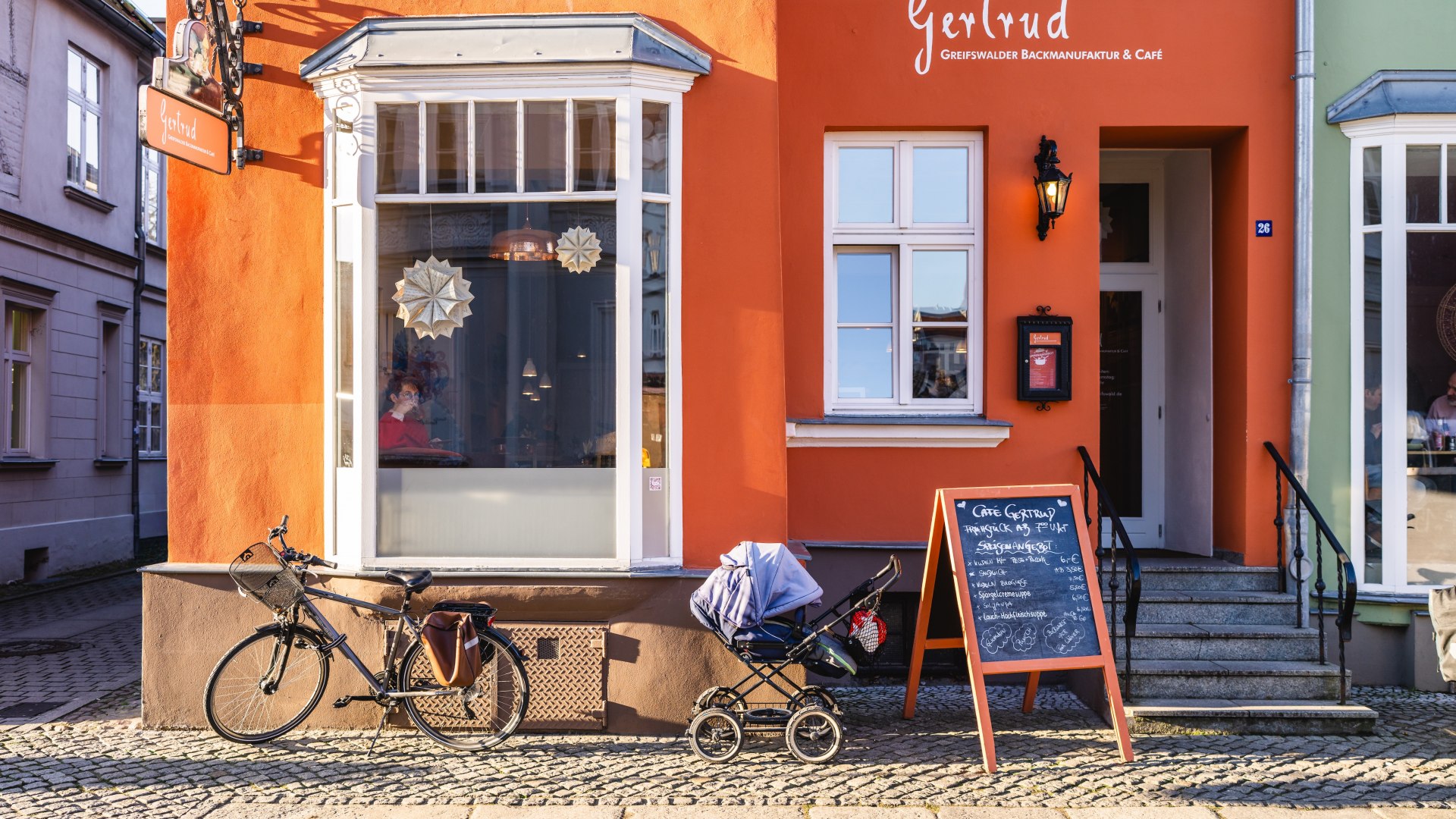 Orangefarbene Fassade des Caf&eacute; Gertrud mit wei&szlig;em Erker in der Greifswalder Altstadt, davor Fahrrad und Kinderwagen.