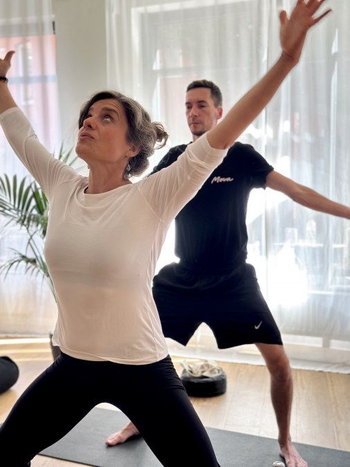 Zwei Personen praktizieren Yoga in einem hellen Studio. Im Vordergrund steht eine Frau in einer kraftvollen stehenden Yoga-Haltung mit erhobenen Armen, im Hintergrund folgt ein Mann derselben Bewegung. Weiches Tageslicht und eine ruhige Atmosph&auml;re unterstreichen Achtsamkeit, Bewegung und K&ouml;rperbewusstsein., &copy; MOVA PLACE - Yoga & Co. Studio