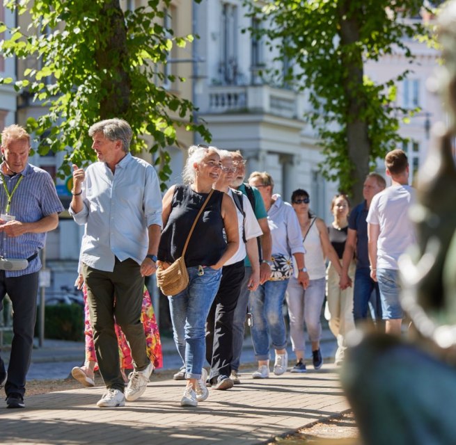Eine Gruppe Menschen ist mit einem Stadtführer auf einem gepflasterten Weg unterwegs. Rechts, unscharf im Bild ist eine Statue am Pfaffenteich zu sehen, im Hintergrund erscheinen die Villen. Blickpunkt sind der Stadtführer sowie ein Paar, dass sich angeregt unterhält. // © Oliver Borchert Eine Gruppe Menschen ist mit einem Stadtführer auf einem gepflasterten Weg unterwegs. Rechts, unscharf im Bild ist eine Statue am Pfaffenteich zu sehen, im Hintergrund erscheinen die Villen. Blickpunkt sind der Stadtführer sowie ein Paar, dass sich angeregt unterhält. // © Oliver Borchert