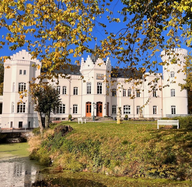 Vor Schloss Schlemmin leuchten Herbstbl&auml;tter &uuml;ber dem Wassergraben, die wei&szlig;e Fassade steht vor blauem Himmel.
