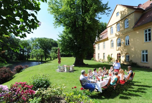 Personen sitzen in einem Garten vor einem Gutshaus mit einer langen Tafel und picknicken bei Sonnenschein. // Kulinarischer Genuss und anregende Gespräche auf Schloss Lühburg // © MV-T/Foto@Andreas-Duerst.de Personen sitzen in einem Garten vor einem Gutshaus mit einer langen Tafel und picknicken bei Sonnenschein.