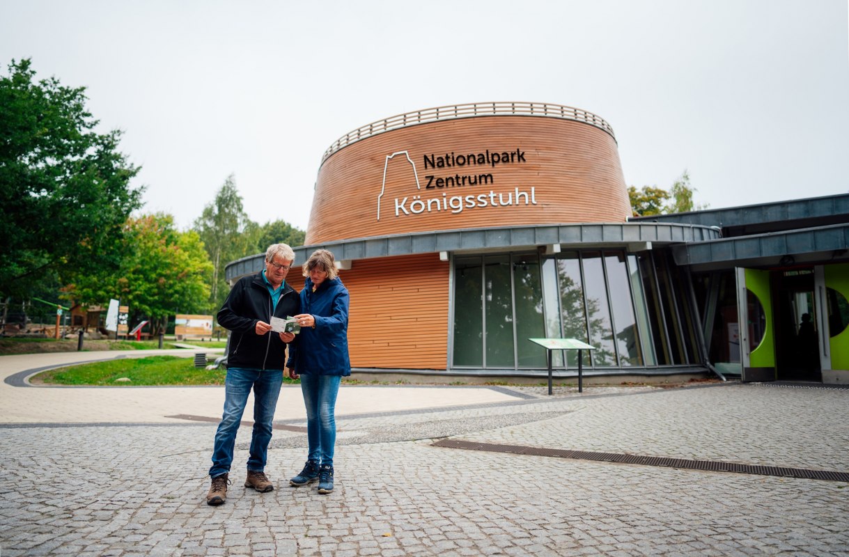 Modernes Gebäude mit rundem Eingangsbereich, vor dem zwei Personen auf einem gepflasterten Platz stehen. // © Nationalpark-Zentrum KÖNIGSSTUHL / Dan Petermann Modernes Gebäude mit rundem Eingangsbereich, vor dem zwei Personen auf einem gepflasterten Platz stehen. // © Nationalpark-Zentrum KÖNIGSSTUHL / Dan Petermann