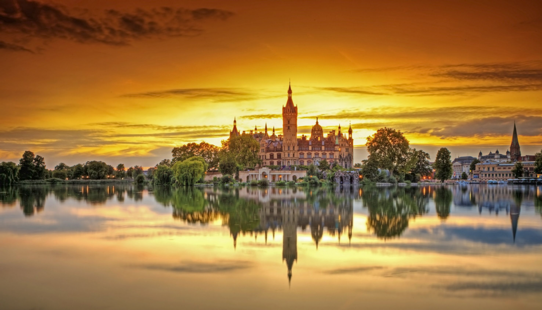 Das Schweriner Schloss wird von den letzten Sonnenstrahlen umrahmt. Der Sonnenuntergang f&auml;rbt den Himmel orange. Im ruhigen See spiegelt sich das Schweriner Schloss.