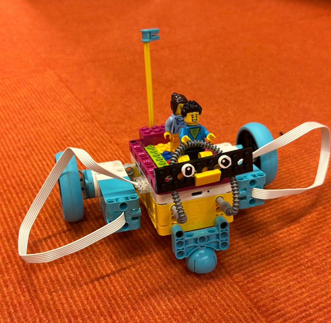 ein kleiner Legoroboter mit zwei Legofiguren darauf, Hintergrund orange // &copy; Stadtbibliothek Stralsund