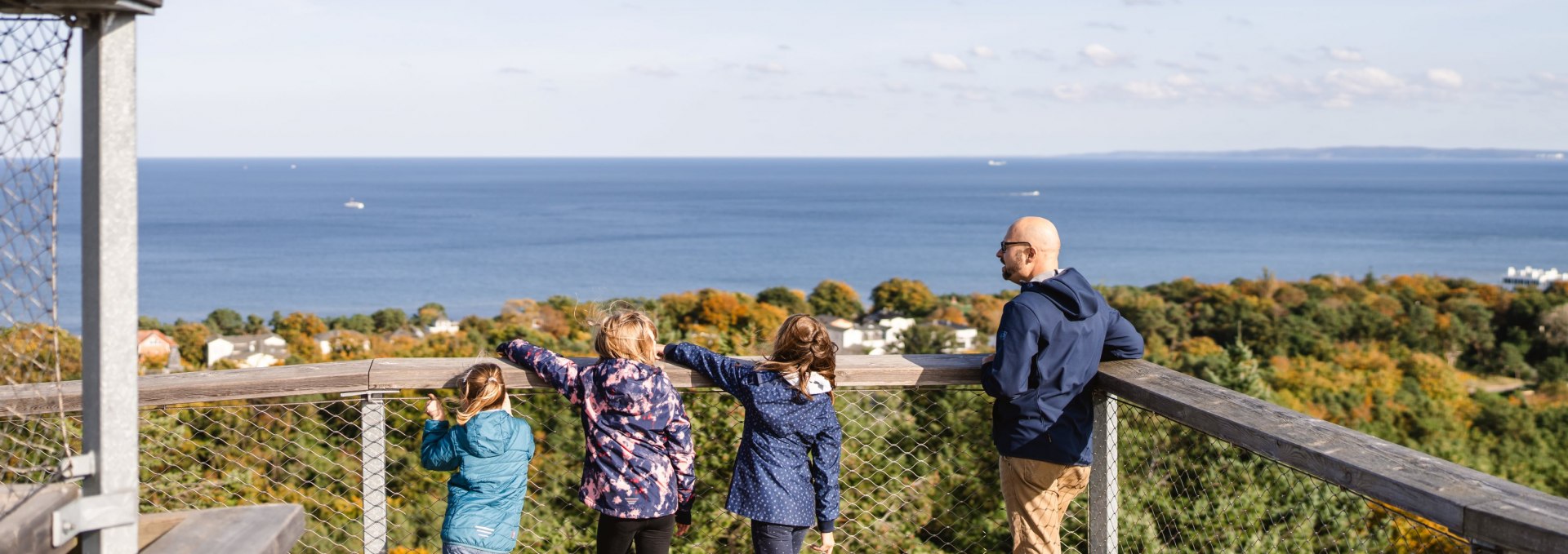  Zwei Erwachsene und drei Kinder stehen auf einer Aussichtsplattform und blicken &uuml;ber den Wald auf das blaue Meer.
