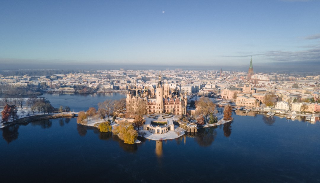 Winterliches Märchenschloss – das Schweriner Schloss auf seiner Insel erstrahlt inmitten verschneiter Landschaft und spiegelndem Wasser als Wahrzeichen der Landeshauptstadt Mecklenburg-Vorpommerns., © TMV/Scholz-Witzel Luftaufnahme des verschneiten Schweriner Schlosses auf einer Insel im See mit Blick auf die Altstadt im Winter.