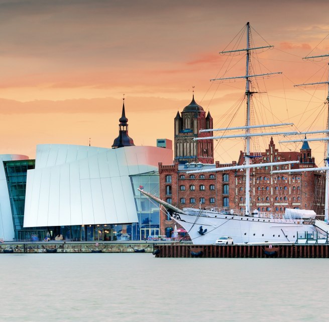 Moderne Museumsarchitektur trifft auf hanseatisches Erbe: Das Ozeaneum am Stralsunder Hafen verschmilzt mit historischen Backsteinbauten und Segelschiffen zu einer einzigartigen Silhouette. Im Abendlicht leuchtet die UNESCO-Welterbestadt in warmen Farbt&ouml;nen. Hier l&auml;sst sich maritime Geschichte hautnah erleben &ndash; vom Museumsdeck bis zur Hafenpromenade. // &copy; Francesco Carovillano
