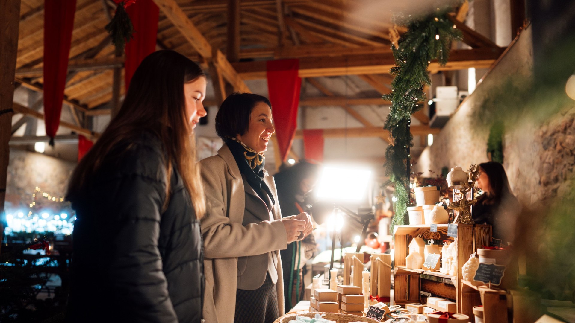 Festliche Stimmung und handgefertigte Schätze, Kerzen und Seifen - einzigartige Weihnachtsgeschenke auf dem Markt., © TMV/Petermann Zwei Frauen stehen an einem weihnachtlich dekorierten Marktstand mit handgemachten Seifen, Kerzen und Geschenkartikeln und genießen die festliche Atmosphäre.