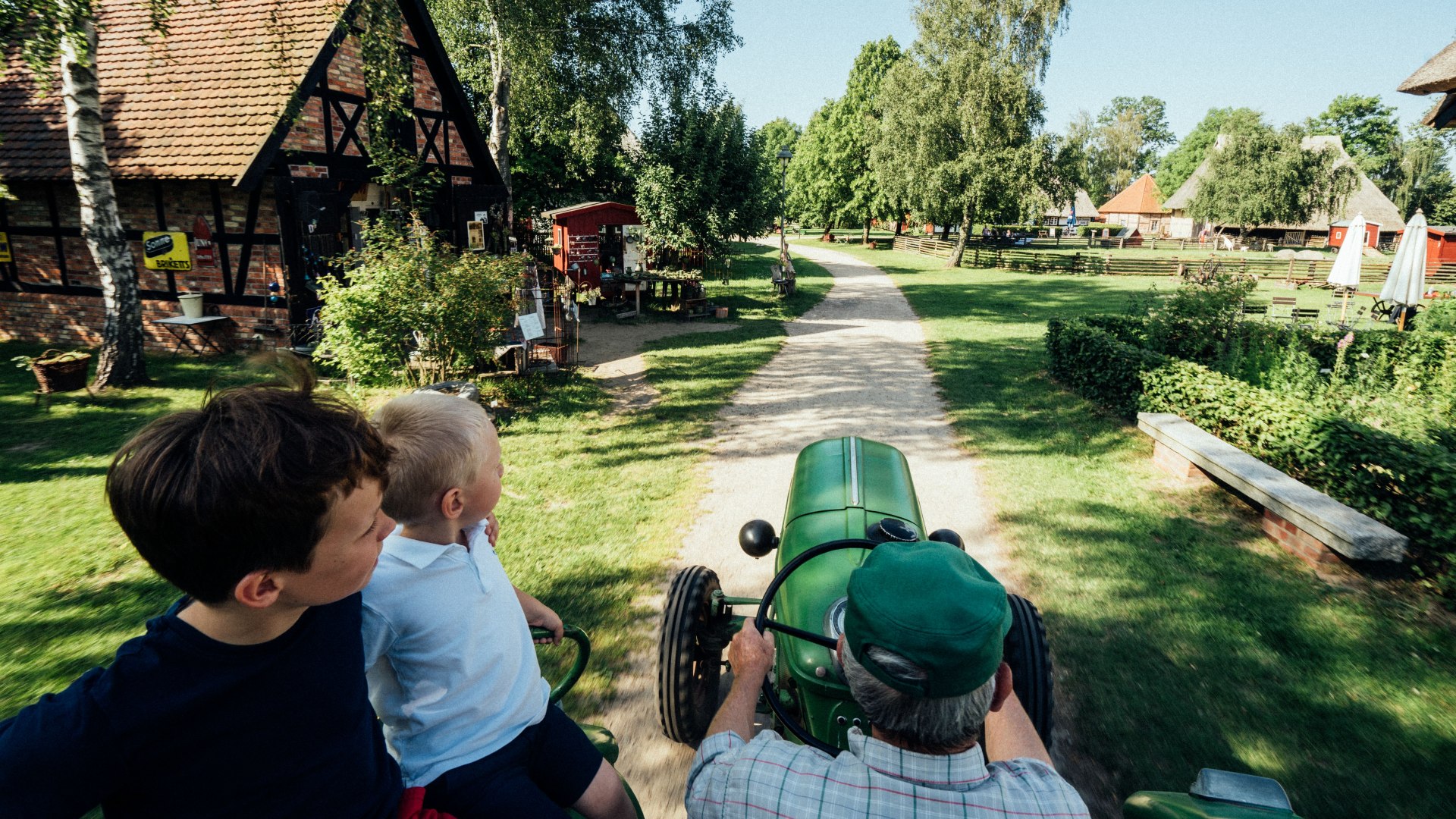 Mit dem historischen Traktor durchs Freilichtmuseum – Familienabenteuer mit ländlichem Charme und lebendiger Geschichte., © TMV/Gänsicke Kinder fahren mit einem älteren Mann auf einem historischen Traktor durch das Freilichtmuseum Klockenhagen mit Fachwerkhäusern und grüner Landschaft.