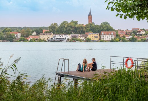 Zwei Frauen sitzen auf einem Steg und blicken dabei auf einen See