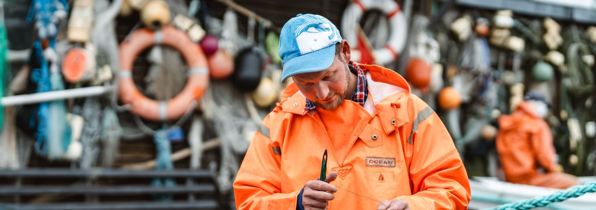 Fischer im Hafen von Gager auf Rügen repariert ein Netz vor einer Hütte mit Rettungsringen und Fischereigeräten, während ein Kollege im Hintergrund sitzt. // Ein Fischer im Hafen von Gager auf Rügen repariert sorgfältig ein Netz. Gekleidet in auffällige orangefarbene Regenkleidung und eine blaue Kappe, steht er vor einer rustikalen Hütte, die mit Rettungsringen und Fischereizubehör dekoriert ist. Im Hintergrund sitzt ein weiterer Fischer, umgeben von der maritimen Atmosphäre des Hafens. // © MV-T/Gross Fischer im Hafen von Gager auf Rügen repariert ein Netz vor einer Hütte mit Rettungsringen und Fischereigeräten, während ein Kollege im Hintergrund sitzt.
