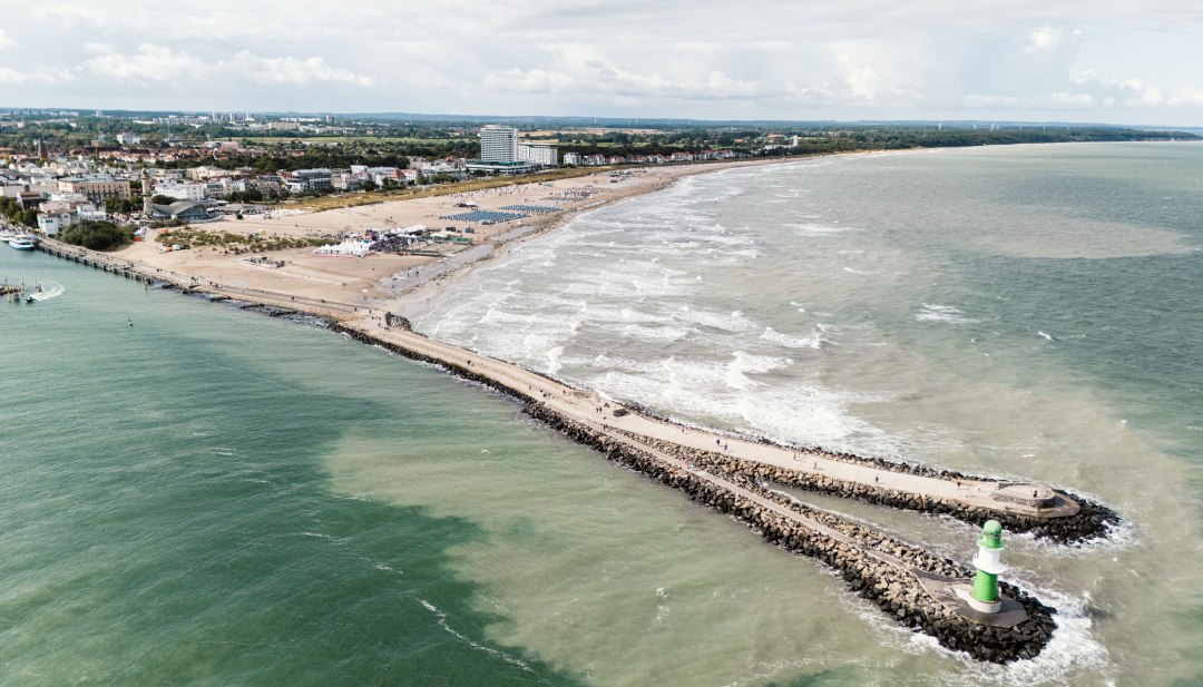 Luftaufnahme vom Strand Warnemünde mit Mole, Leuchtturm und Ostsee bei sanftem Wellengang. // Die Ostsee legt sich in sanften Wellen an den breiten Sandstrand von Warnemünde. Die steinerne Mole führt hinaus aufs Meer, gekrönt vom grün-weißen Leuchtturm. Strandkörbe reihen sich entlang der Küste, während sich dahinter die Silhouette des Seebads mit historischen Gebäuden und modernen Hotels abzeichnet. // © MV-T/Scholz-Witzel Luftaufnahme vom Strand Warnemünde mit Mole, Leuchtturm und Ostsee bei sanftem Wellengang.