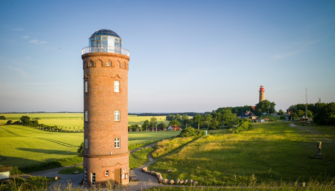 Schinkelturm aus rotem Backstein und Leuchtturm am Kap Arkona auf der Insel R&uuml;gen, umgeben von gr&uuml;nen Wiesen und Feldern, mit Blick auf die Ostsee im Hintergrund.