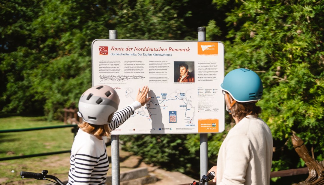 Zwei Radfahrende deuten auf eine Infotafel mit Karte, auf der die Route der Norddeutschen Romantik verläuft. // In der Dorfkirche Kemnitz wurde Friedrich August von Klinkowström getauft. Die Route der Norddeutschen Romantik führt von hier über Schloss Ludwigsburg bis nach Wolgast. // © MV-T/Gross Zwei Radfahrende deuten auf eine Infotafel mit Karte, auf der die Route der Norddeutschen Romantik verläuft.