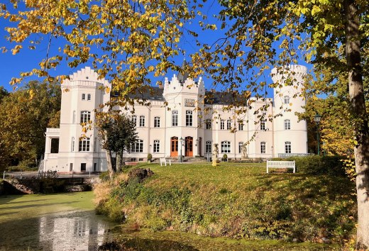 Vor Schloss Schlemmin leuchten Herbstbl&auml;tter &uuml;ber dem Wassergraben, die wei&szlig;e Fassade steht vor blauem Himmel.