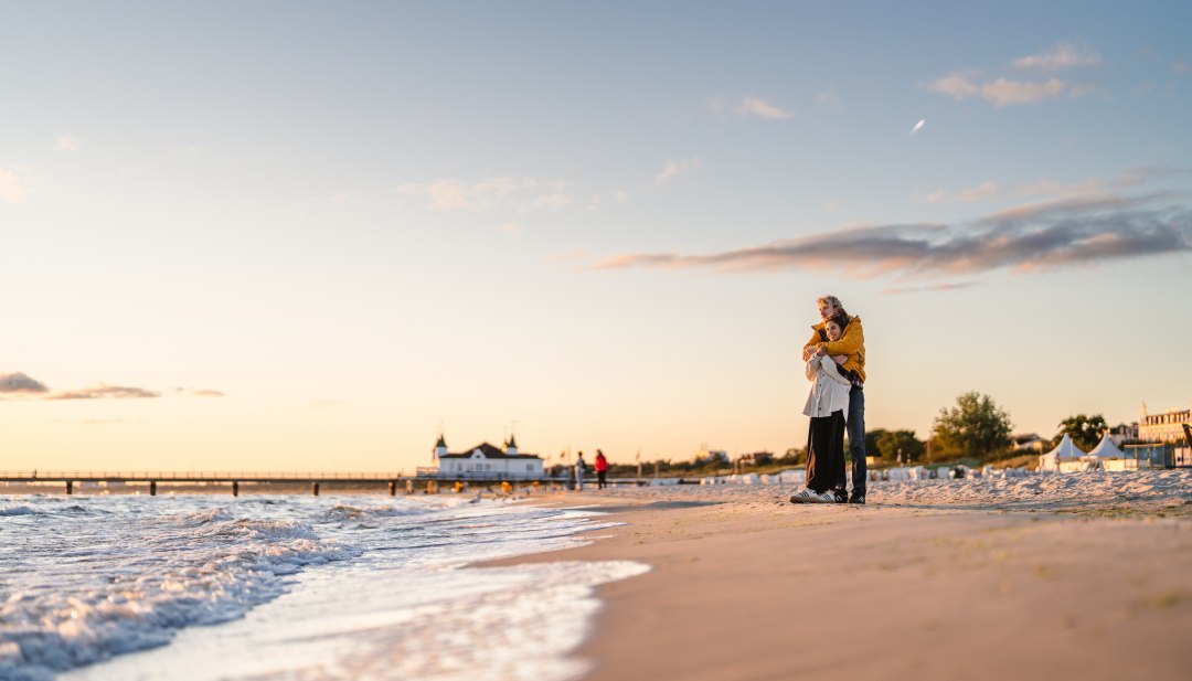 Ein Paar steht am Strand von Ahlbeck in Umarmung, die Seebrücke und Abendhimmel im warmen Licht. // Sonnenuntergang am Strand von Ahlbeck: gemeinsam am Wasser entlanggehen, die Weite spüren, kurz loslassen. // © MV-T/Gross Ein Paar steht am Strand von Ahlbeck in Umarmung, die Seebrücke und Abendhimmel im warmen Licht.