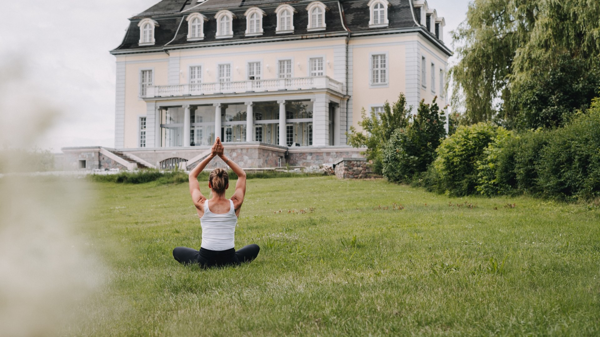 Frau meditiert auf einer Wiese vor dem historischen Herrenhaus "The Reset Hotel" mit weißen Säulen. // Meditationshaltung vor dem The Reset Hotel. // © Alessandro Schleiss Frau meditiert auf einer Wiese vor dem historischen Herrenhaus "The Reset Hotel" mit weißen Säulen.