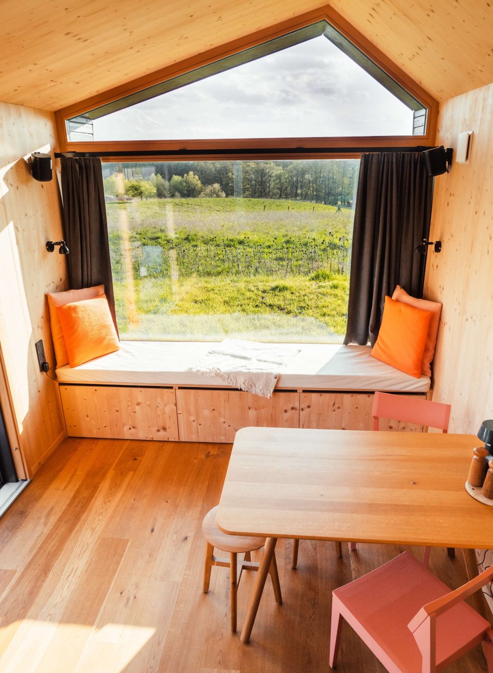Stilvoller Minimalismus trifft auf Natur – Tiny House mit Panoramafenster und gemütlichem Interior für eine entspannte Auszeit., © mari.linni || Nicola Link Berlin Helles Tiny House mit großer Fensterfront, Holzinterieur und Sitzbank mit Blick auf eine weite, grüne Landschaft.