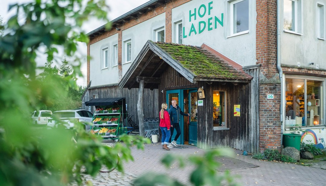 Rustikaler Hofladen mit begrüntem Dach, frischem Obst- und Gemüsestand draußen und zwei Personen am Eingang. // Regionale Köstlichkeiten direkt vom Erzeuger – ein Besuch im idyllischen Hofladen mit frischen Produkten und ländlichem Charme. // © MV-T/Gross Rustikaler Hofladen mit begrüntem Dach, frischem Obst- und Gemüsestand draußen und zwei Personen am Eingang.