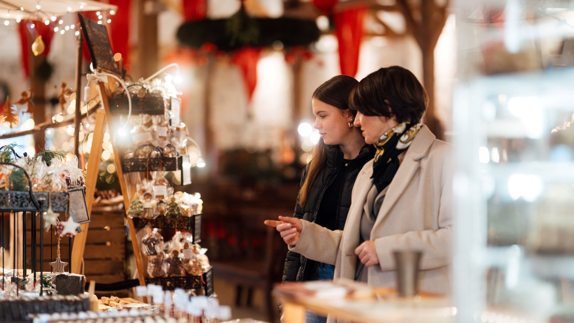Zwei Frauen schlendern über einen stimmungsvoll beleuchteten Weihnachtsmarkt und betrachten kunstvoll verpackte Süßigkeiten und Dekorationen. // Handgemachte Leckereien und Geschenke auf dem Weihnachtsmarkt. // © MV-T/Petermann Zwei Frauen schlendern über einen stimmungsvoll beleuchteten Weihnachtsmarkt und betrachten kunstvoll verpackte Süßigkeiten und Dekorationen.