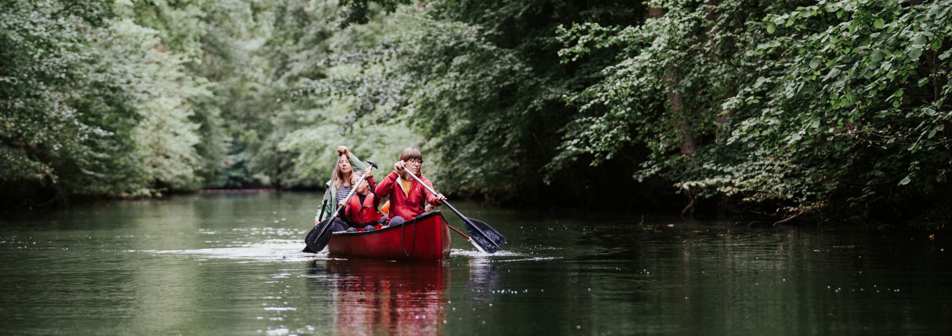 Zwei Personen paddeln im roten Kanu durch einen von Bäumen gesäumten Fluss im Müritz-Nationalpark.