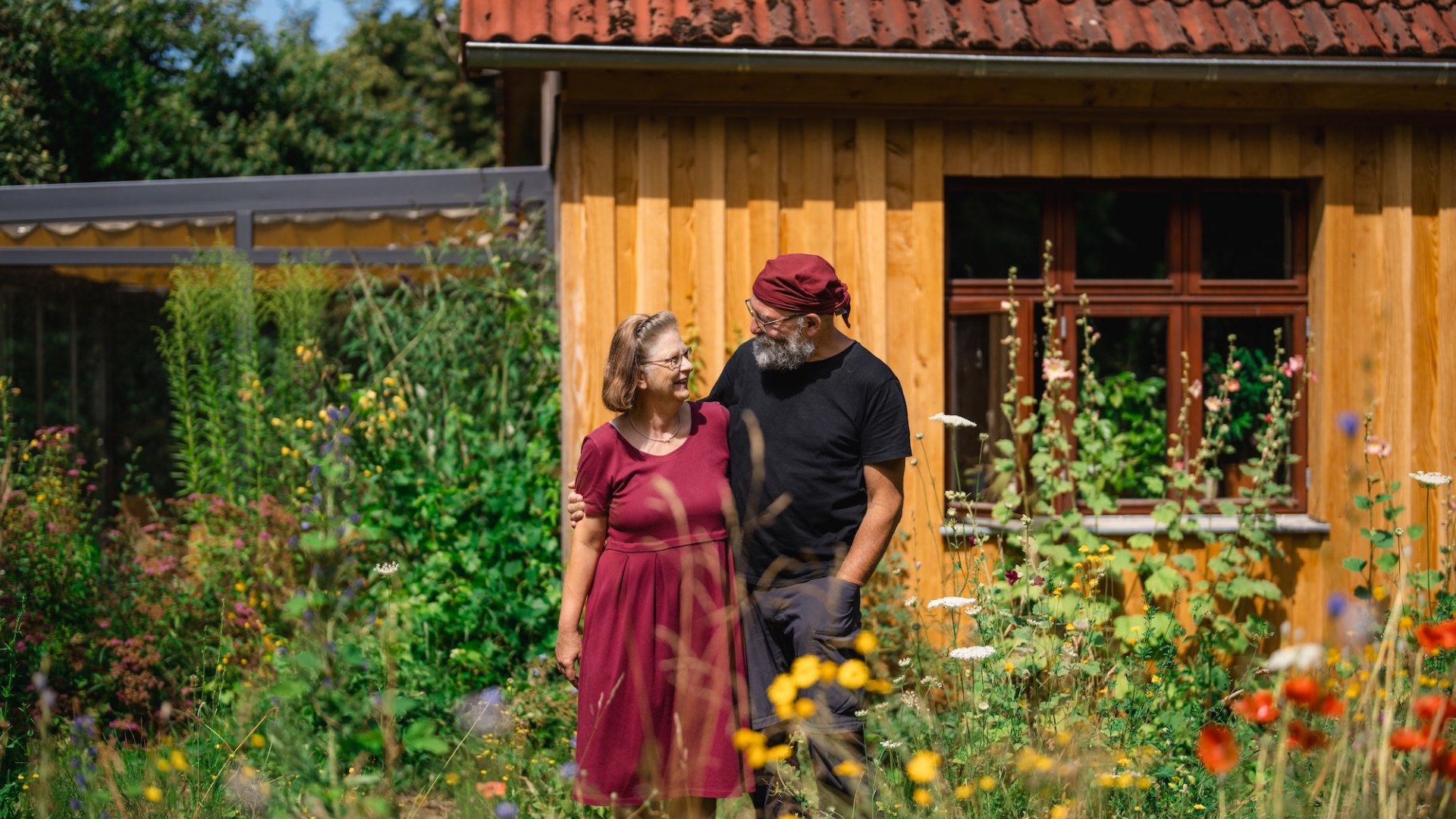 Et par st&aring;r i en blomstrende have foran et tr&aelig;hus og smiler til hinanden, med farvestr&aring;lende vilde blomster i forgrunden.