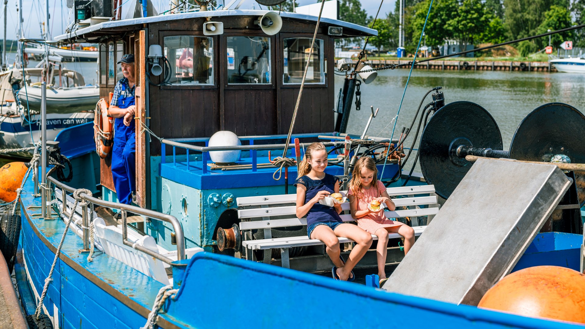 Zwei Kinder essen ein Fischbrötchen auf einem Fischkutter, der normaler Weise zum Fischen rausfährt. Der Kapitän steht in der Tür des Fahrerhäuschens. // Leckere Fischbrötchen im Hafen von Karlshagen. Und den besten Platz direkt auf dem Kutter. // © MV-T/Tiemann Zwei Kinder essen ein Fischbrötchen auf einem Fischkutter, der normaler Weise zum Fischen rausfährt. Der Kapitän steht in der Tür des Fahrerhäuschens.
