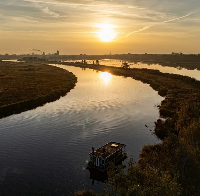 Hausboot auf der Peene bei Sonnenuntergang, umgeben von ruhiger Flusslandschaft und goldenem Licht am Horizont.