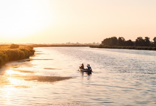 Zwei Personen paddeln in einem Kanu &uuml;ber einen ruhigen Fluss bei Sonnenuntergang, mit goldenem Licht auf dem Wasser.