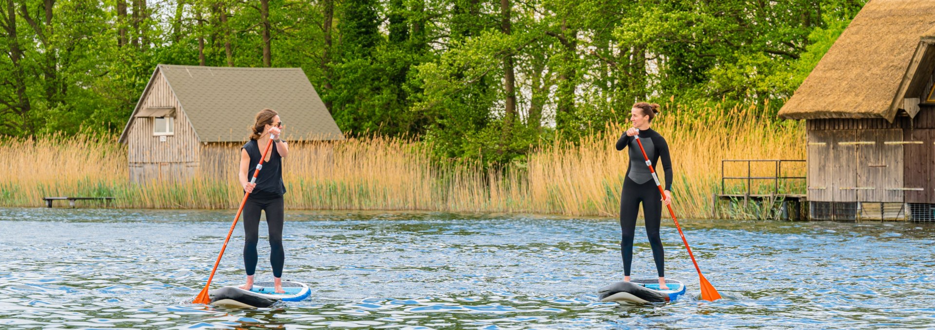 Zwei Frauen beim Stand-up-Paddling vor reetgedeckten Bootsh&auml;usern und dichten Schilfg&uuml;rteln in der Mecklenburgischen Seenplatte.