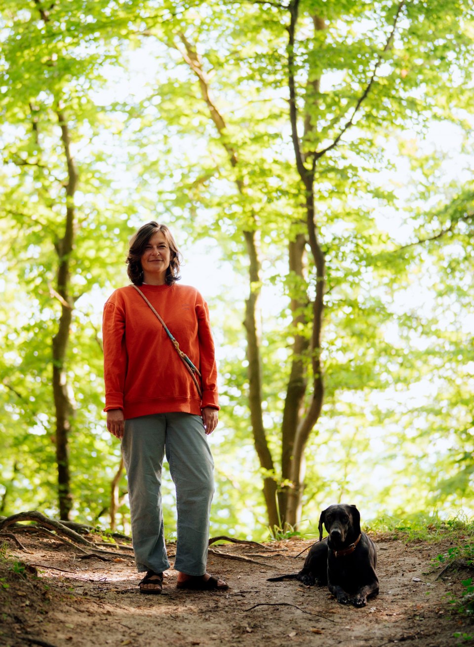 Eine Frau steht auf einem Waldweg in der Granitz auf Rügen, neben ihr liegt ein schwarzer Hund im Schatten. // Anne mit ihrer Hündin Mascha. // © MV-T/Petermann Eine Frau steht auf einem Waldweg in der Granitz auf Rügen, neben ihr liegt ein schwarzer Hund im Schatten.