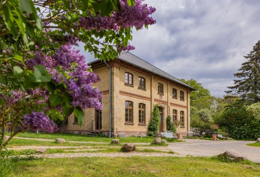 Gutshaus Lexow mit blühendem Flieder im Vordergrund und historischer Backsteinfassade. // Violette Fliederblüten rahmen das Gutshaus Lexow ein, während sich der Himmel über der historischen Fassade auftürmt. Zwischen Grün und Blütenpracht atmet der Ort Ruhe – ein Platz zum Ankommen und Durchatmen. // © Gutshaus Lexow Gutshaus Lexow mit blühendem Flieder im Vordergrund und historischer Backsteinfassade.