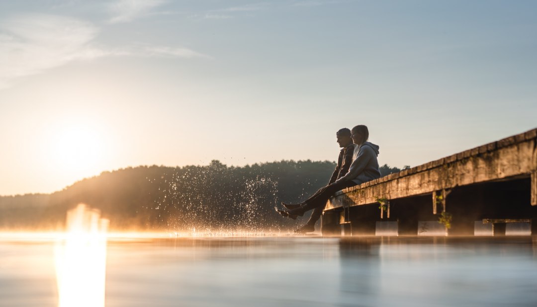 Zwei Personen sitzen im Sonnenaufgang auf einem Steg am Mirower See und lassen die F&uuml;&szlig;e ins glatte Wasser tauchen, w&auml;hrend feiner Nebel &uuml;ber der Oberfl&auml;che schwebt.