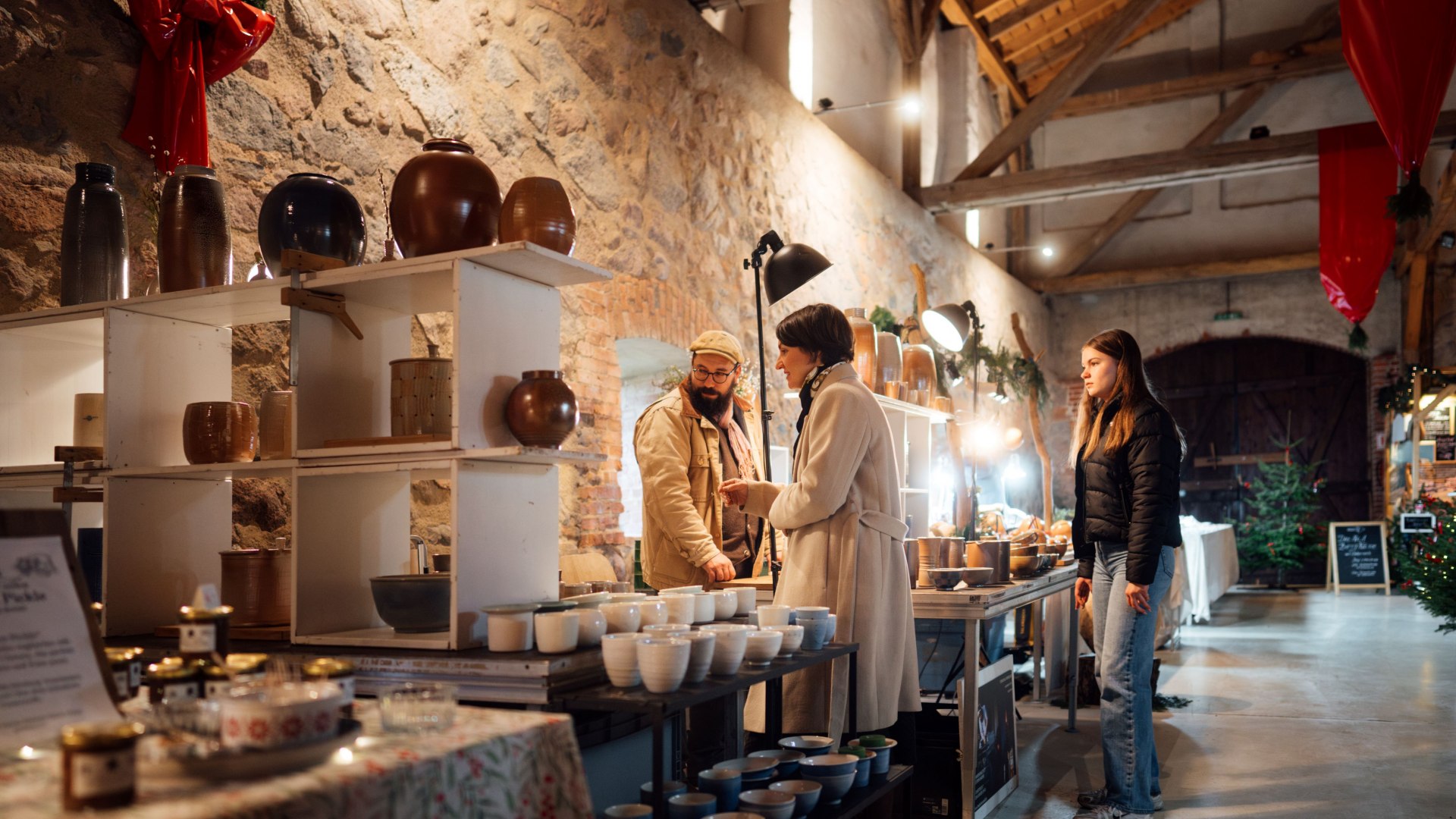 Ein Verkäufer auf einem Weihnachtsmarkt präsentiert handgefertigte Keramikschalen und Vasen, während eine Besucherin interessiert nachfragt. // Handgemachte Keramik und stilvolle Unikate – ein Marktstand voller einzigartiger Geschenkideen für die Weihnachtszeit. // © MV-T/Petermann Ein Verkäufer auf einem Weihnachtsmarkt präsentiert handgefertigte Keramikschalen und Vasen, während eine Besucherin interessiert nachfragt.