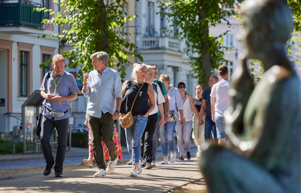 Eine Gruppe Menschen ist mit einem Stadtführer auf einem gepflasterten Weg unterwegs. Rechts, unscharf im Bild ist eine Statue am Pfaffenteich zu sehen, im Hintergrund erscheinen die Villen. Blickpunkt sind der Stadtführer sowie ein Paar, dass sich angeregt unterhält., © Oliver Borchert