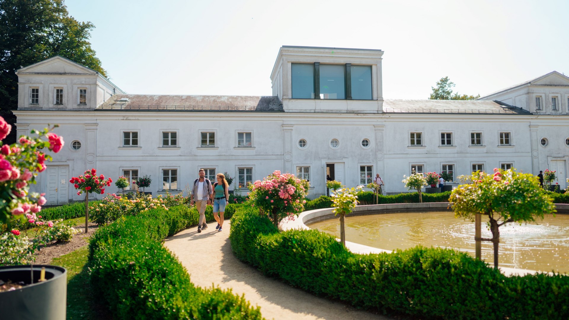 Ein Paar schlendert entlang eines gepflegten Weges durch den Garten der Orangerie in Putbus. Der Garten ist mit Rosen und Buchsbaumhecken geschmückt, während im Vordergrund ein kleiner Teich mit Springbrunnen sichtbar ist. Im Hintergrund steht das historische Gebäude der Orangerie mit weißen Fassaden und großen Fenstern. // Ein Spaziergang durch den blühenden Garten der Orangerie in Putbus auf Rügen – ein Ort voller Eleganz und romantischer Atmosphäre. // © MV-T/Petermann Ein Paar schlendert entlang eines gepflegten Weges durch den Garten der Orangerie in Putbus. Der Garten ist mit Rosen und Buchsbaumhecken geschmückt, während im Vordergrund ein kleiner Teich mit Springbrunnen sichtbar ist. Im Hintergrund steht das historische Gebäude der Orangerie mit weißen Fassaden und großen Fenstern.