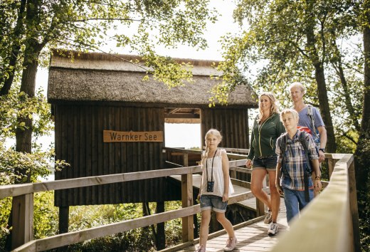 Eine Familie geht über einen Holzsteg zu einer Hütte mit Reetdach, ein Mädchen trägt ein Fernglas um den Hals. // Knarrende Holzbohlen, raschelndes Laub: Am Warnker See im Müritz-Nationalpark erkunden Familien den Steg zur Reetdachhütte. // © MV-T/Roth Eine Familie geht über einen Holzsteg zu einer Hütte mit Reetdach, ein Mädchen trägt ein Fernglas um den Hals.