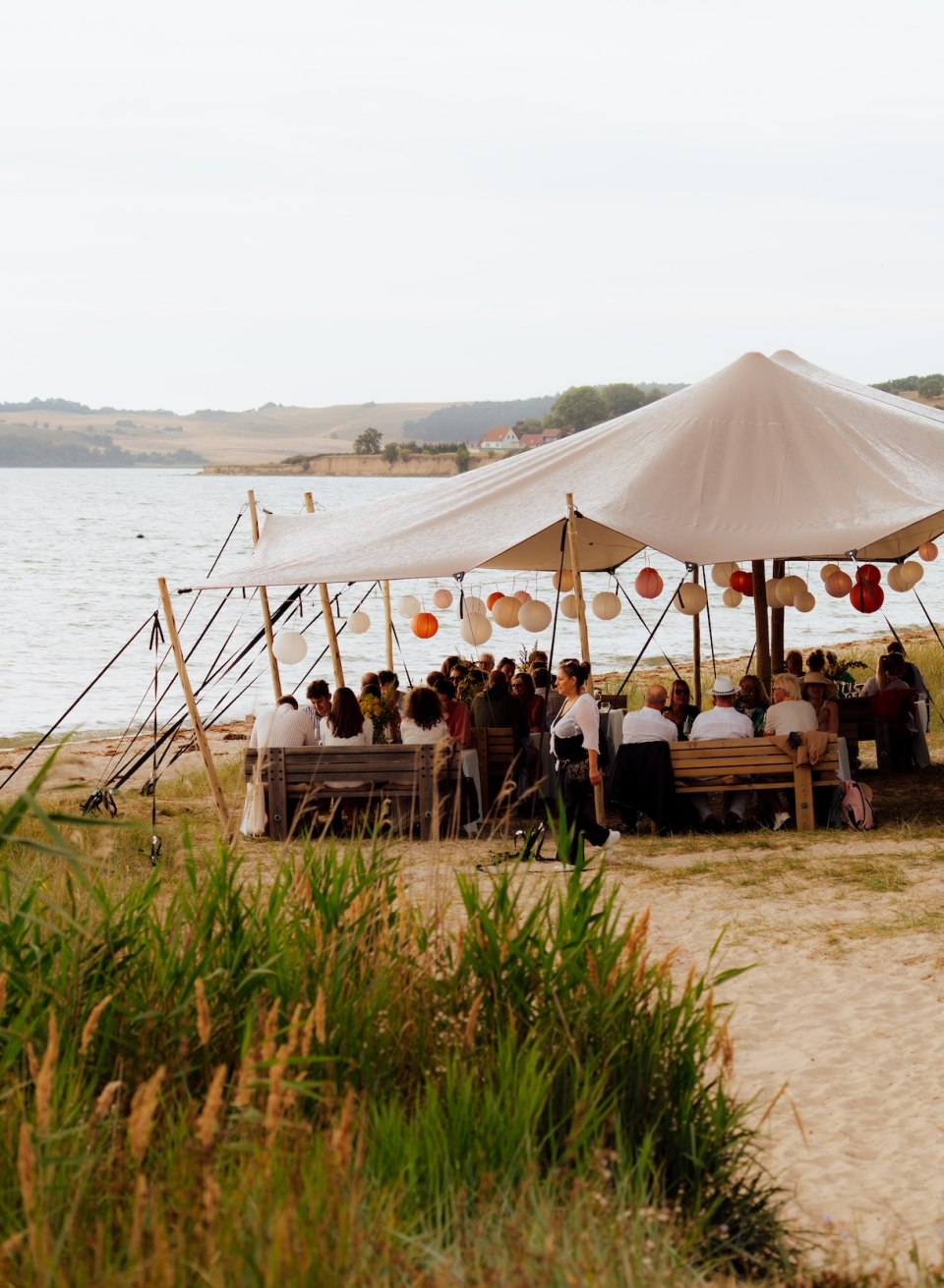 Gäste sitzen an langen Tafeln unter einem Zelt im Dünengras am Strand von Thiessow, daneben liegt das Meer. // Unter dem Zeltdach im Dünengras kommt die Runde zusammen – Dinner am Strand von Thiessow, mit Blick aufs Wasser. // © MV-T/Petermann Gäste sitzen an langen Tafeln unter einem Zelt im Dünengras am Strand von Thiessow, daneben liegt das Meer.