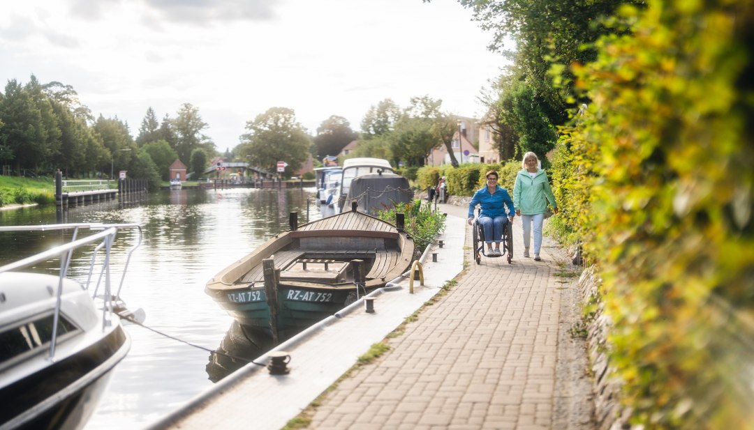 Eine Rollstuhlfahrerin unterwegs mit Ihrer Freundin am Kanal in Plau am See. An der Kante liegen Boote.