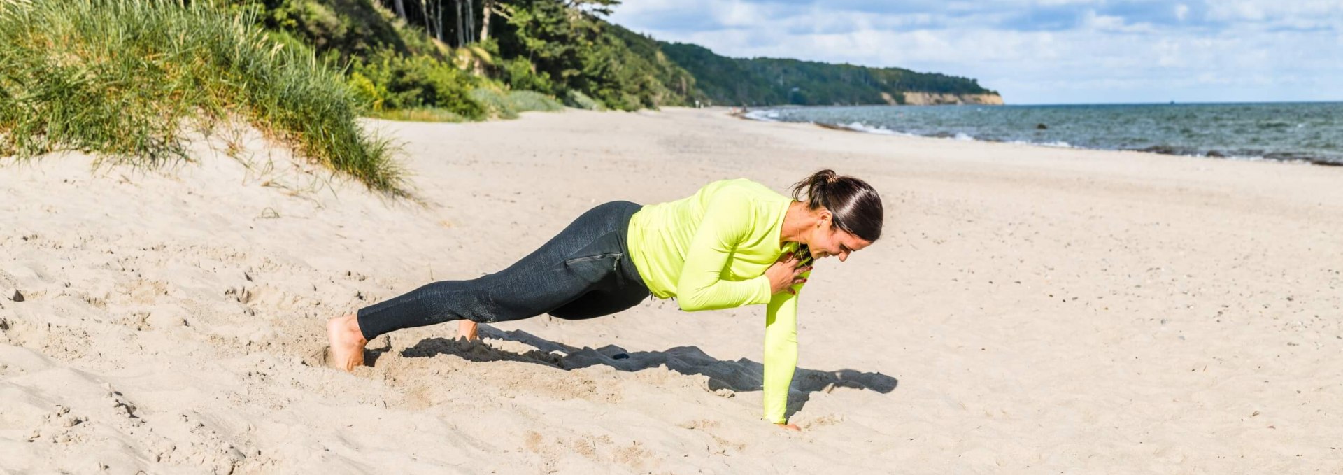 Stütz dich auf die Handflächen. Wenn du einen stabilen Stand hast, hebst du die linke Hand vom Boden ab und führst sie zur rechten Schulter. Einmal antippen und dann zurück auf den Strandsand. Achte darauf, dass Hüften und Körper nicht zur Seite schwingen, wenn du dir auf die Schultern tippst. Es soll sich ausschließlich die Hand zur gegenüberliegenden Schulter bewegen. 6-10 Wiederholungen pro Seite, © TMV/Tiemann Eine Frau ist am Strand und macht Fitnessübungen: hier Plank mit Schulter-Tippen