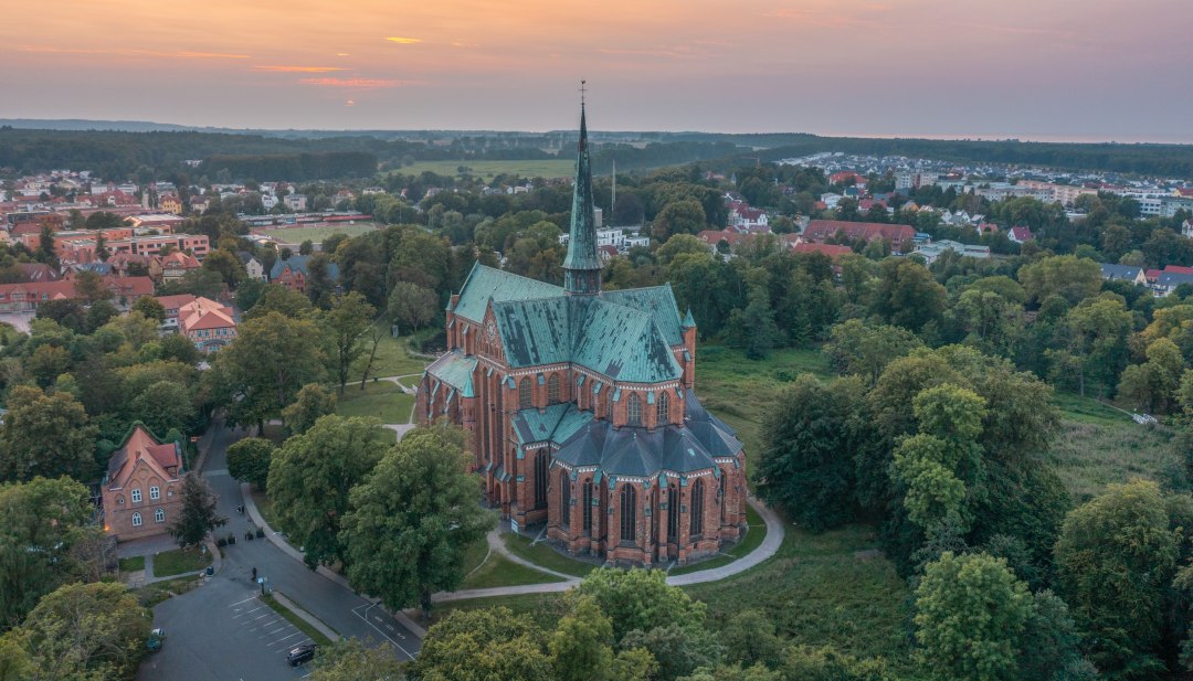 Das beeindruckende Münster in Bad Doberan – ein Meisterwerk der Backsteingotik inmitten idyllischer Natur und historischer Stadtlandschaft., © TMV/Nordombord Luftaufnahme des Münsters in Bad Doberan bei Sonnenuntergang, umgeben von Bäumen, Wiesen und der umliegenden Stadt.