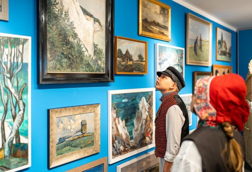 Zeichnungen und Malereien h&auml;ngen an der Wand im Heimatmuseum M&ouml;nchgut - Kinder stehen in traditioneller Kleidung davor und begutachten.